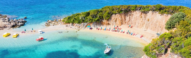 Luftaufnahme des Ksamil Strandes in Albanien, mit klarem türkisfarbenem Wasser, bunten Sonnenschirmen und Liegestühlen. Kleine Boote sind am Ufer verankert, und üppiges Grün umrahmt den Strand.