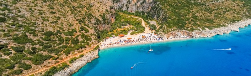 Luftaufnahme des Gjipe Strandes in Albanien, mit klarem türkisfarbenem Wasser und einer langen Straße, die entlang der Küste führt. Der Strand ist von beeindruckenden Klippen und üppigem Grün umgeben, was eine abgeschiedene und malerische Kulisse schafft.