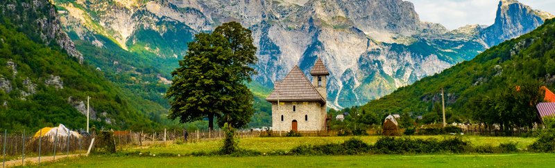 Die Kirche von Theth in Albanien, umgeben von den imposanten Bergen der albanischen Alpen. Die malerische Kirche mit ihrem grauen Ziegeldach steht in einem grünen Tal und ist ein historisches Wahrzeichen des Dorfes Theth. Im Hintergrund erheben sich steile Felswände und üppige Wälder, die die Schönheit und Abgeschiedenheit dieser Region betonen.